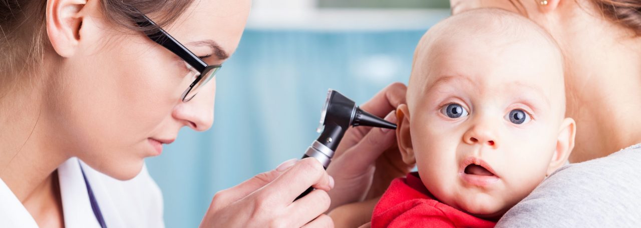 Photo of an audiologist using an otoscope to examine a child held by another adult